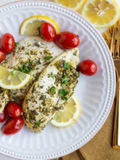Two chicken breasts on a white plate with cherry tomatoes, sliced lemons and golden napkin