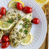 Two chicken breasts on a white plate with cherry tomatoes, sliced lemons and golden napkin