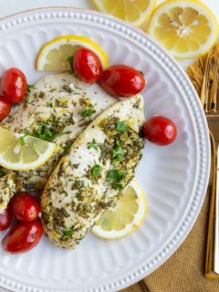 Two chicken breasts on a white plate with cherry tomatoes, sliced lemons and golden napkin