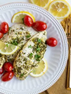 Two chicken breasts on a white plate with cherry tomatoes, sliced lemons and golden napkin