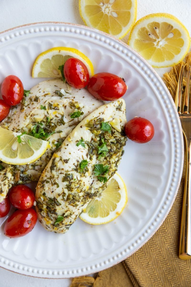 Two chicken breasts on a white plate with cherry tomatoes, sliced lemons and golden napkin