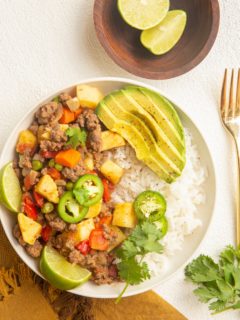 Bowl of Mexican Picadillo with rice and avocado. Limes, fork, and napkin off to the side.