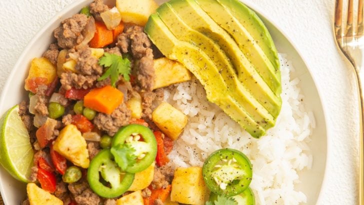 Bowl of Mexican Picadillo with rice and avocado. Limes, fork, and napkin off to the side.