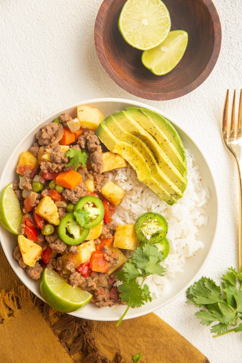 Bowl of Mexican Picadillo with rice and avocado. Limes, fork, and napkin off to the side.