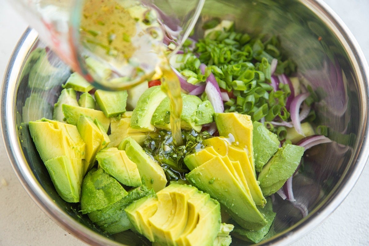 Ingredients for avocado salad in a stainless steel mixing bowl with the dressing being poured in.