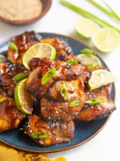 angle shot of blue plate of chicken thighs. limes and green onion in the background.