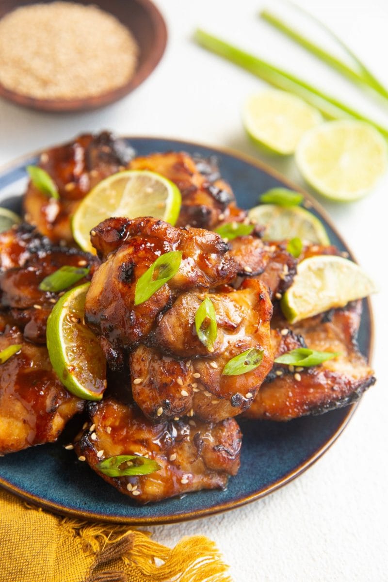 angle shot of blue plate of chicken thighs. limes and green onion in the background.