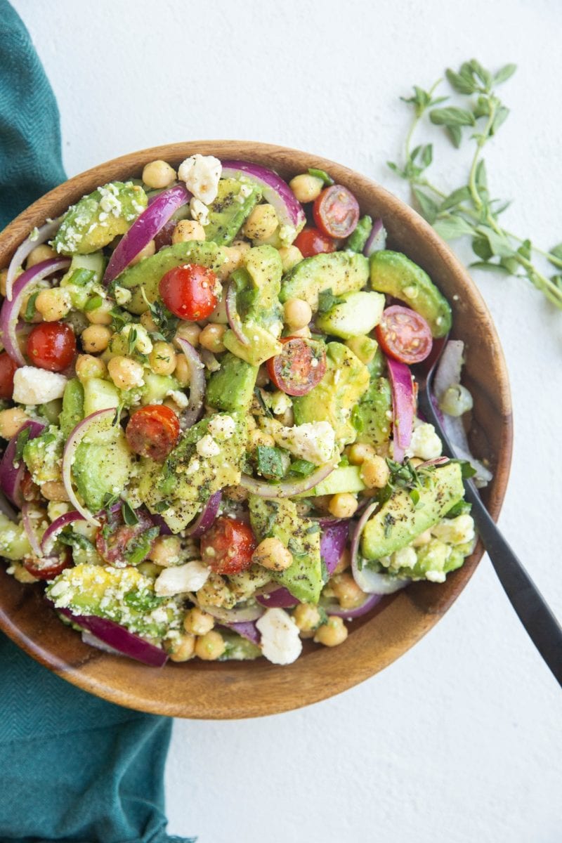 Top down photo of a wooden bowl of chickpea avocado salad with fresh oregano to the side and a blue napkin