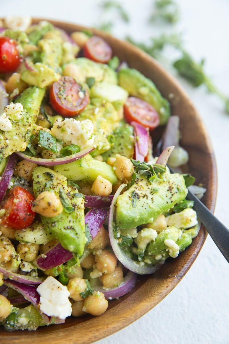 Wooden bowl full of garbanzo beans, avocado, red onion, green onion, feta cheese, cherry tomatoes and lemon herb dressing.