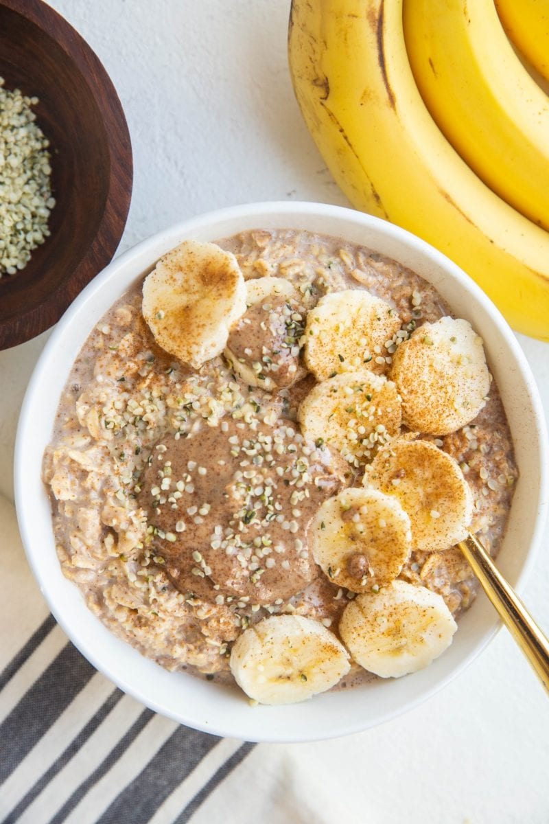 White bowl of overnight oatmeal with sliced bananas, a sprinkle of cinnamon, almond butter and hemp seeds. With fresh bananas to the side, a napkin, gold spoon and bowl of hemp seeds