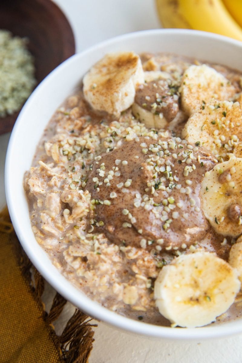 Close up of a bowl of oatmeal with a dollop of almond butter, cinnamon, fresh bananas, and hemp seeds on top.