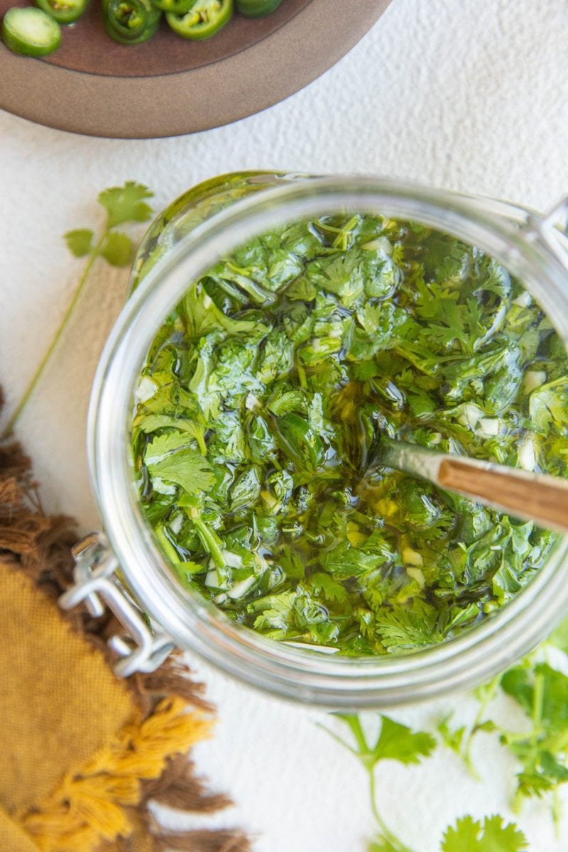 Top down photo of a jar of chimichurri sauce with a plate of sliced serranoes, cilantro and a napkin to the side.