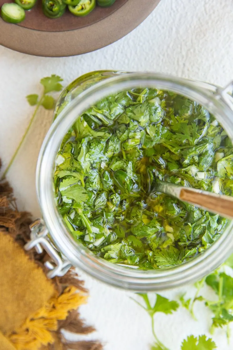 Top down photo of a jar of chimichurri sauce with a plate of sliced serranoes, cilantro and a napkin to the side.