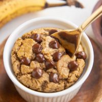 Ramekin of baked oats with chocolate chips on top and a golden spoon with ripe bananas in the background