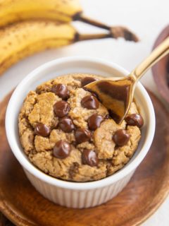 Ramekin of baked oats with chocolate chips on top and a golden spoon with ripe bananas in the background