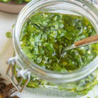 Jar of chimichurri sauce with a napkin to the side, along with fresh cilantro and jalapenos in the background.