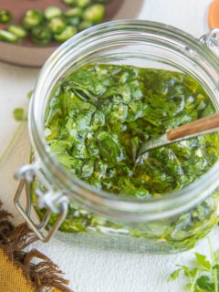 Jar of chimichurri sauce with a napkin to the side, along with fresh cilantro and jalapenos in the background.