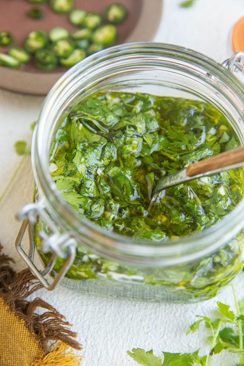 Jar of chimichurri sauce with a napkin to the side, along with fresh cilantro and jalapenos in the background.