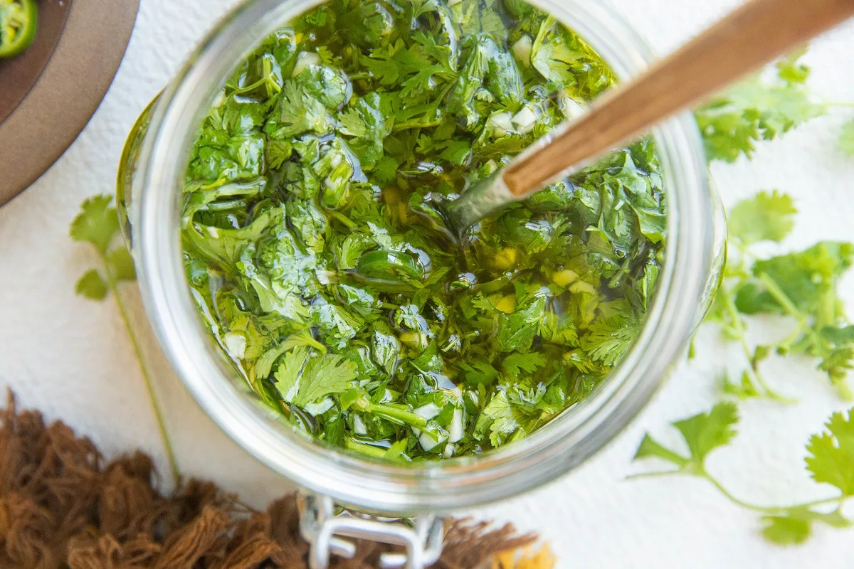 horizontal image of chimichurri sauce in a jar with a spoon in it and fresh herbs to the side.
