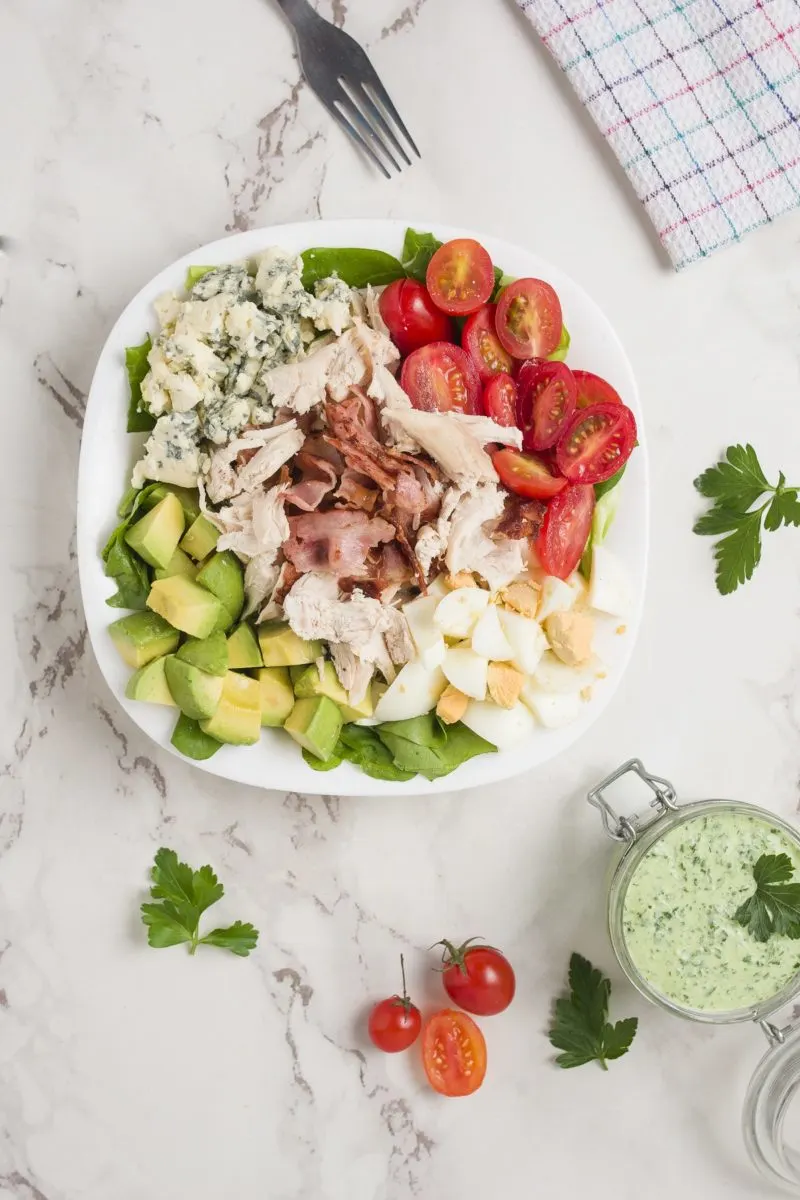 Top down photo of Cobb Salad on a white surface with a napkin and dressing to the side.