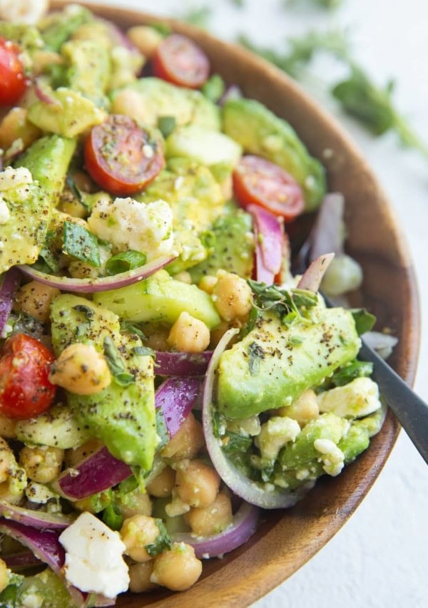 Wooden bowl full of garbanzo beans, avocado, red onion, green onion, feta cheese, cherry tomatoes and lemon herb dressing.
