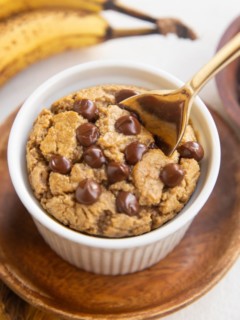 Ramekin of baked oats with chocolate chips on top and a golden spoon with ripe bananas in the background