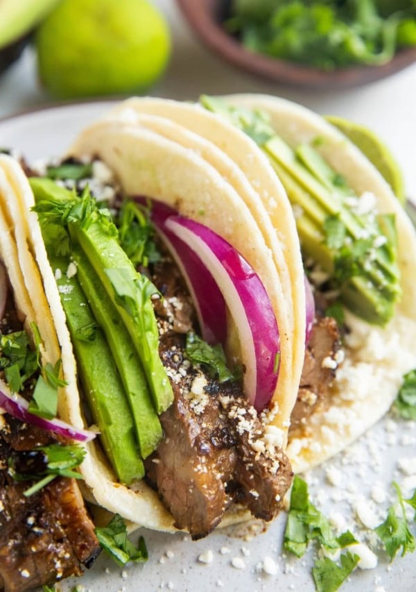 Flank steak tacos on a plate with fresh cilantro, lime, and avocado in the background