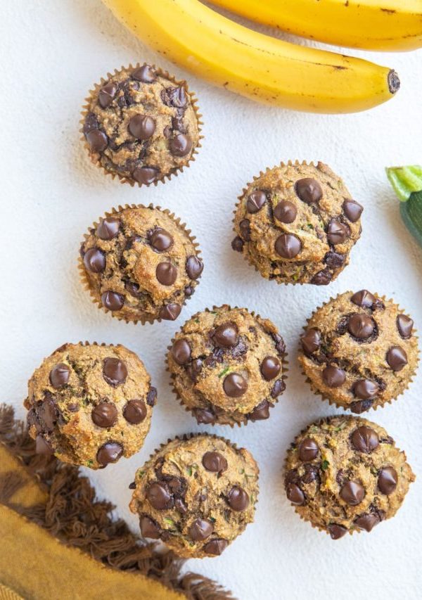Top down photo of a whole batch of banana zucchini muffins on a white backdrop with bananas and a zucchini squash to the side, along with a golden napkin.