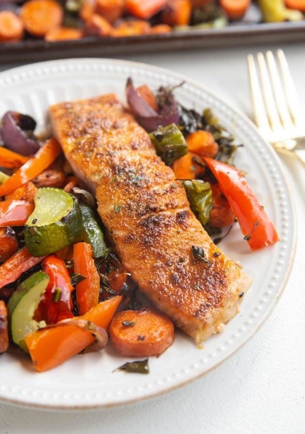 White plate with a salmon filet on it and roasted veggies next to it. Sheet pan of veggies in the background.