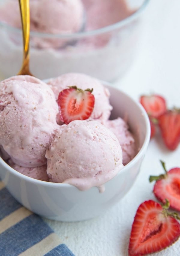 White bowl of strawberry ice cream with a gold spoon and fresh strawberries to the side.
