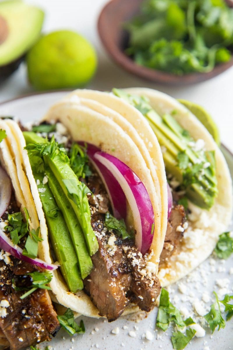 Flank steak tacos on a plate with fresh cilantro, lime, and avocado in the background