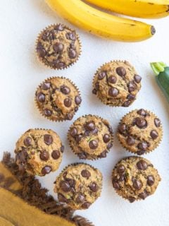 Top down photo of a whole batch of banana zucchini muffins on a white backdrop with bananas and a zucchini squash to the side, along with a golden napkin.