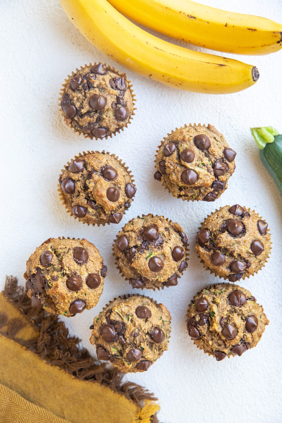 Top down photo of a whole batch of banana zucchini muffins on a white backdrop with bananas and a zucchini squash to the side, along with a golden napkin.