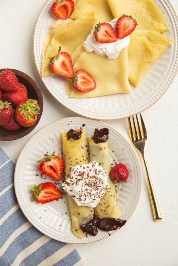 Two plates of crepes, one plate has chocolate filled crepes with strawberries.