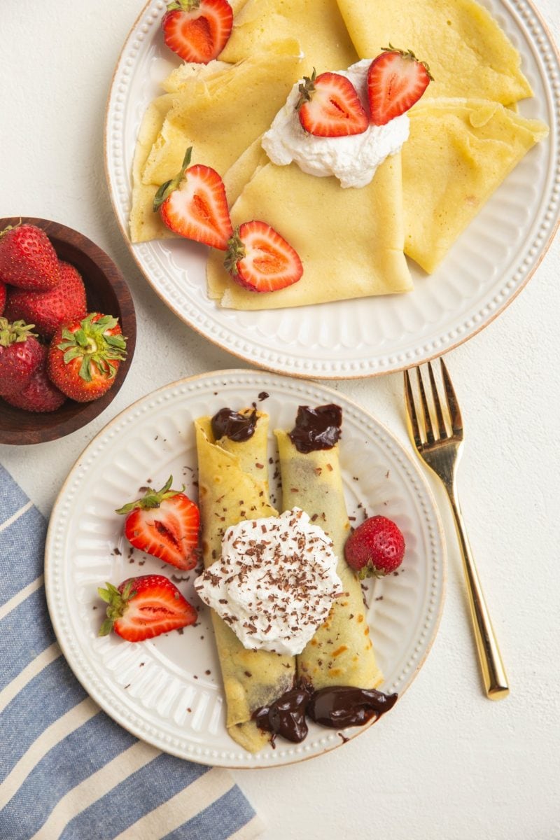 Two plates of crepes, one plate has chocolate filled crepes with strawberries.