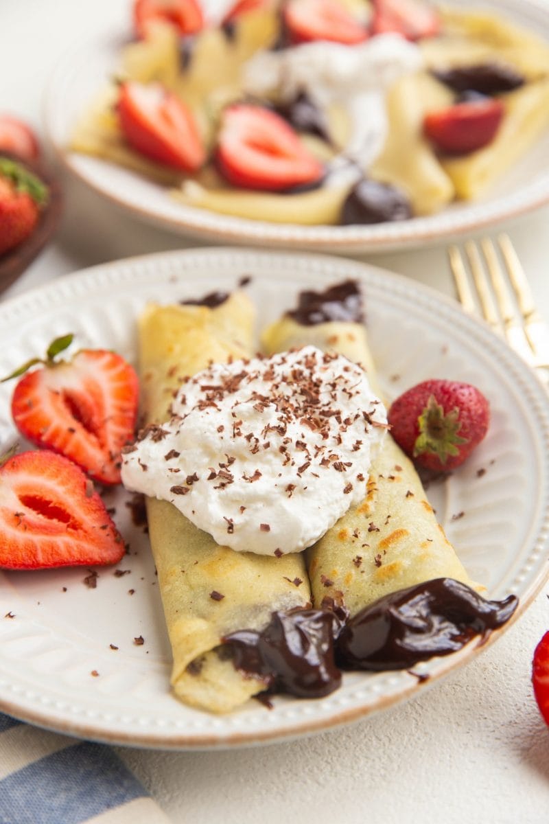 Two crepes on a white plate filled with ganache, topped with whipped cream, chocolate shavings and fresh strawberries.
