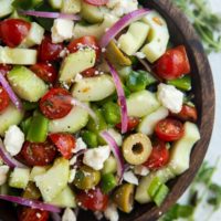Wooden bowl of traditional Greek Salad with fresh herbs to the side.