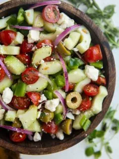 Wooden bowl of traditional Greek Salad with fresh herbs to the side.