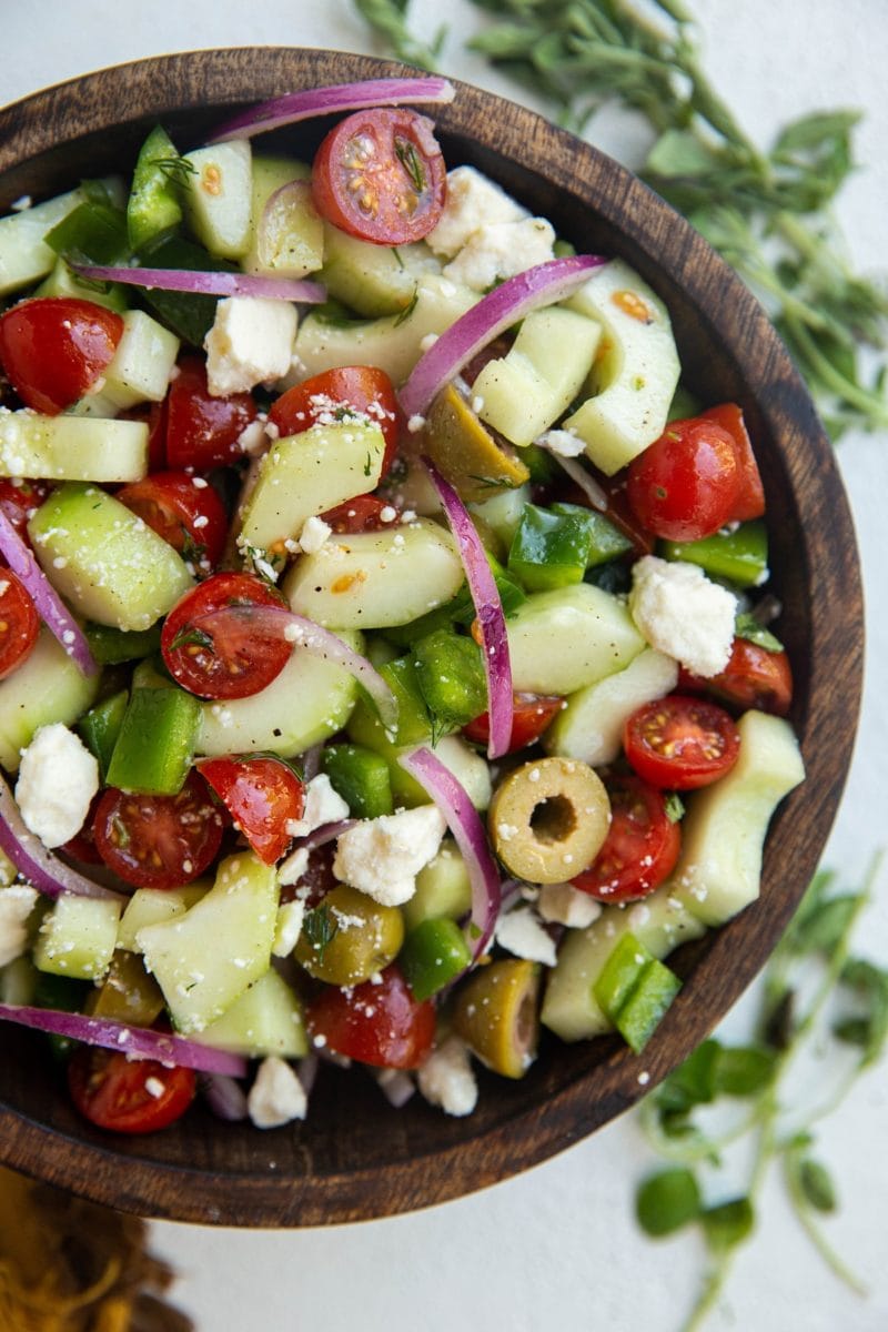 Wooden bowl of traditional Greek Salad with fresh herbs to the side.