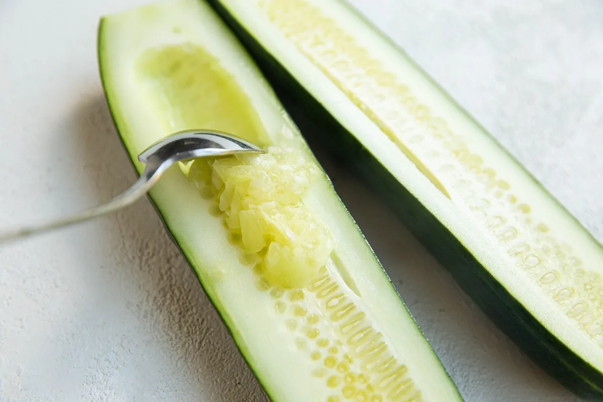 Raw cucumber sitting on a cutting board sliced in half with a spoon scooping out the seeds.