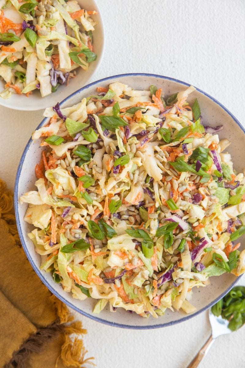 Large bowl of healthy colelsaw and a small white bowl of coleslaw with a napkin and spoon to the side.