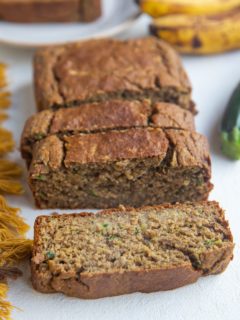 Loaf of zucchini banana bread sliced on a white backdrop with a plate with a slice on it in the back.