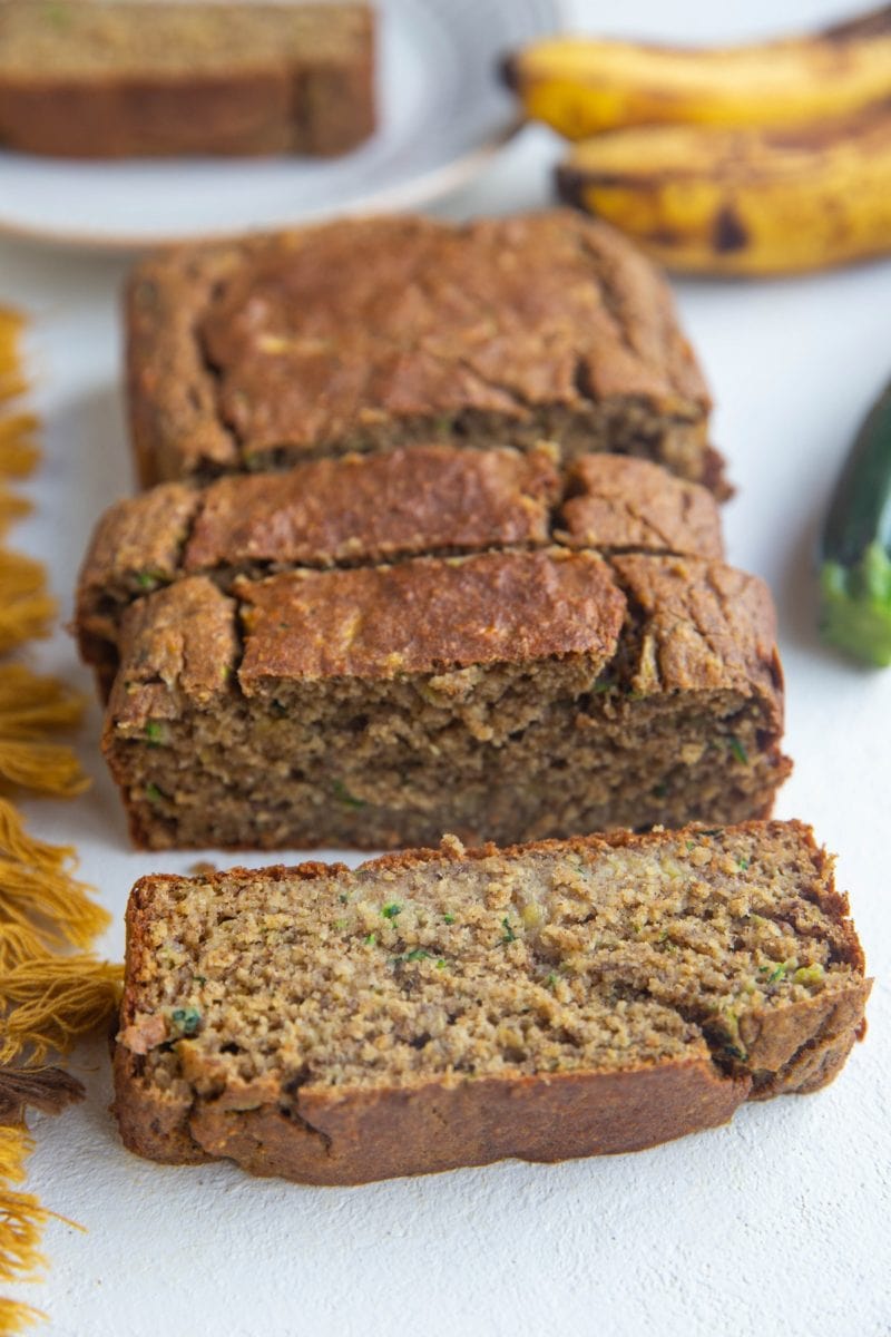 Loaf of zucchini banana bread sliced on a white backdrop with a plate with a slice on it in the back.