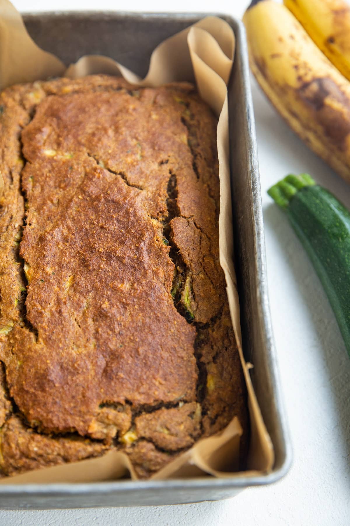 Loaf of zucchini banana bread in a loaf pan with a fresh zucchini and ripe bananas in the background.