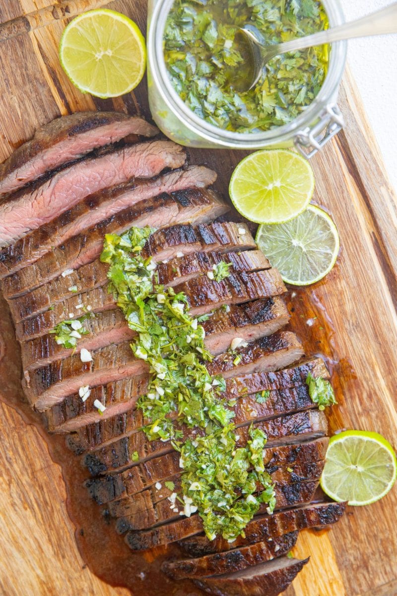 wooden cutting board with sliced steak on top with a jar of chimichurri sauce to the side with sliced limes.