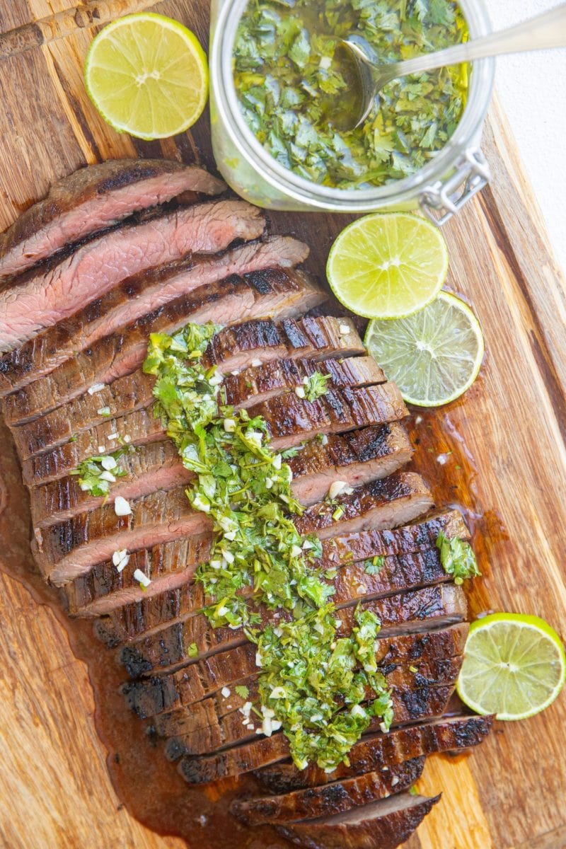 wooden cutting board with sliced steak on top with a jar of chimichurri sauce to the side with sliced limes.