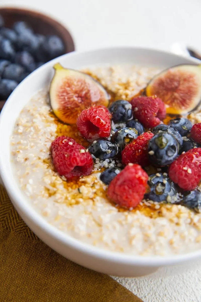 close up image of steel cut oatmeal with fresh berries, figs, and honey on top with a golden napkin and a bowl of blueberries in the background.