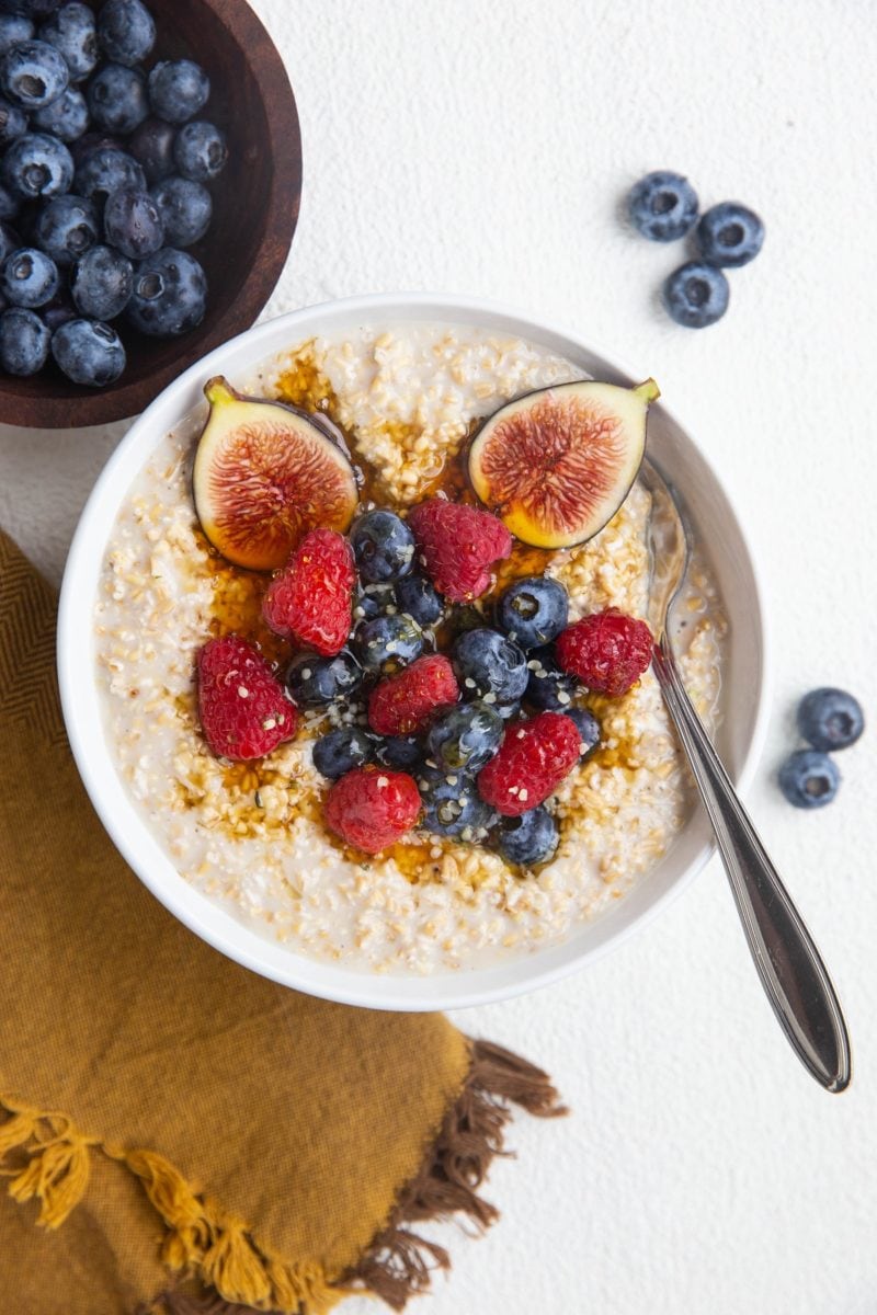 White bowl of steel cut oatmeal, loaded with fresh fruit with a silver spoon and a bowl of blueberries to the side.