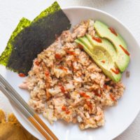 White bowl of salmon and rice mixed together with avocado and seaweed snacks on the side.