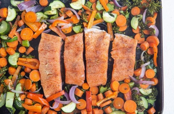 Sheet pan of partially roasted veggies with raw salmon in the middle, ready to be roasted